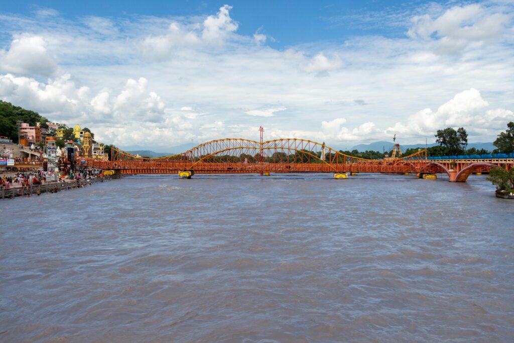 A vibrant rusty bridge over a bustling river with a backdrop of cloudy skies and distant hills.